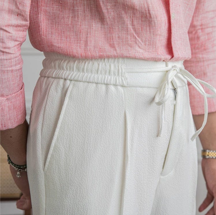 Man wearing white linen tailored pants with drawstring waist, standing on patterned rug indoors.